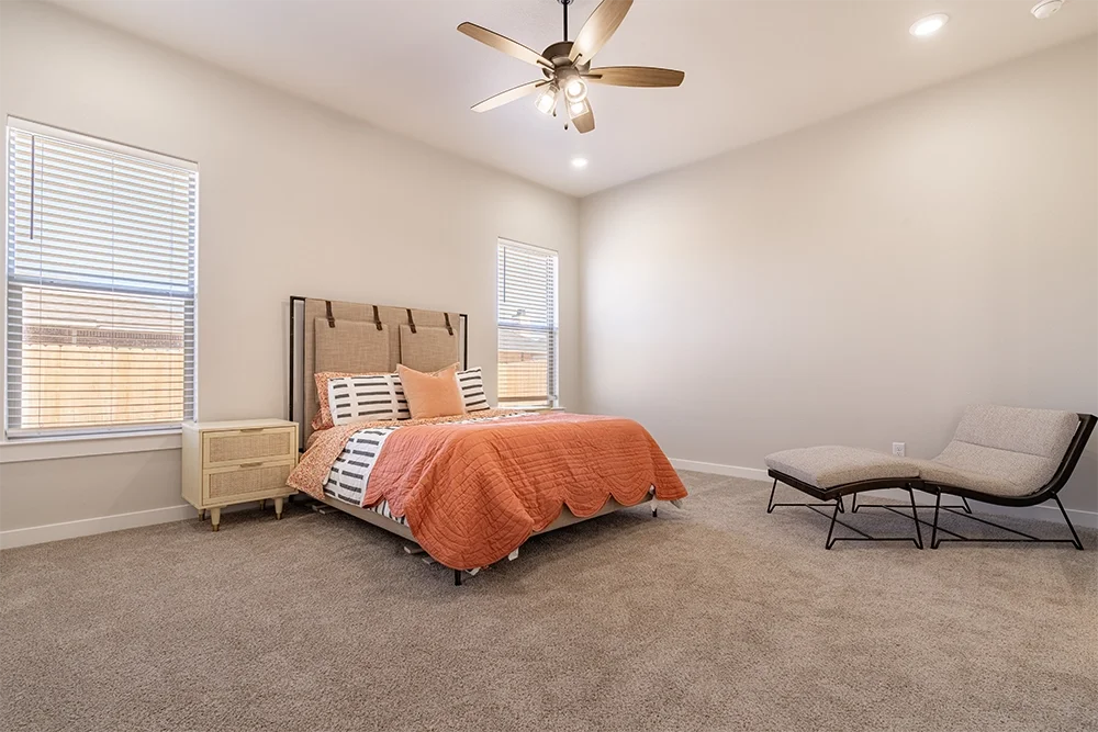 A bedroom with a bed that has a beige headboard, orange comforter, and striped pillows. There are two windows with blinds, a beige nightstand, and a modern lounge chair with a small matching ottoman. Ceiling fan with lights and carpeted floor.