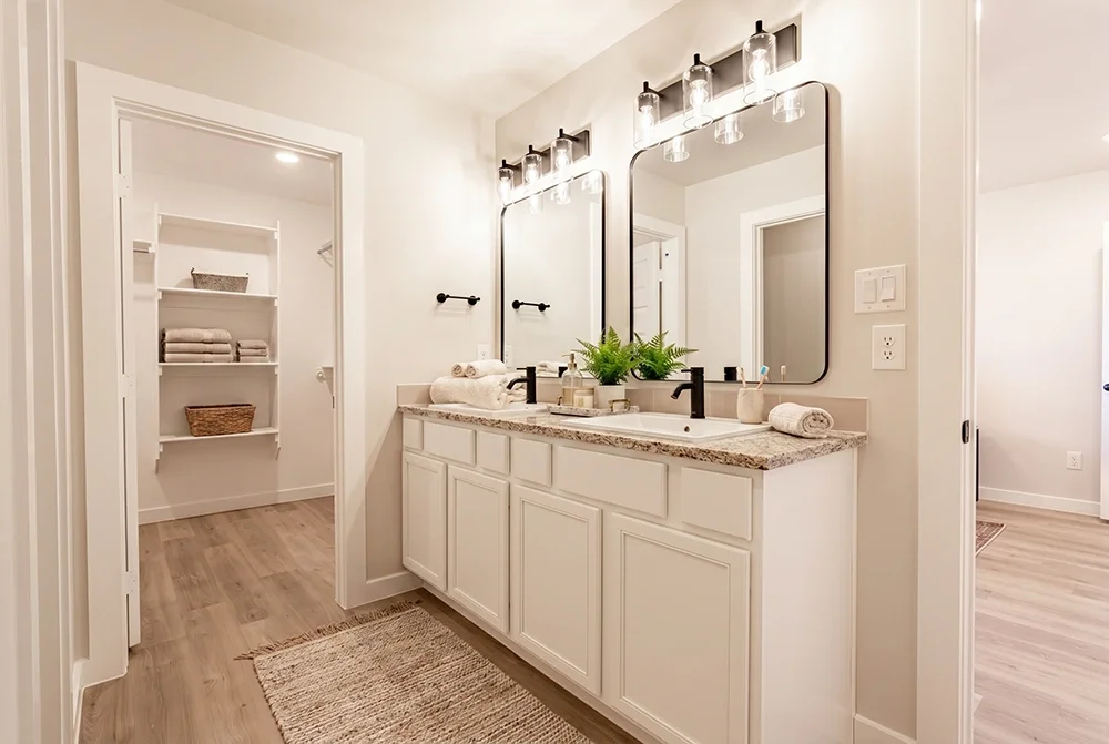Modern bathroom with double vanity, granite countertop, large mirrors, and pendant lights, with a walk-in closet visible through an open door.