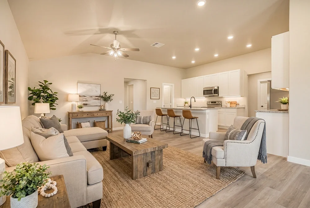 Open-concept living room and kitchen with neutral decor, beige sofa, armchair, wooden coffee table, kitchen island with barstools, modern appliances, and plants.