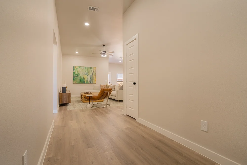 Empty hallway leading into a living room with beige walls, wooden floor, and minimal decor.