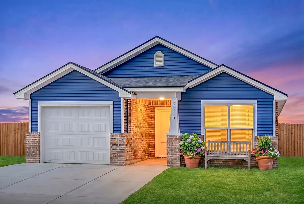 A blue house with a white garage door, brick accents, and front porch with a bench and flower pots, during twilight.