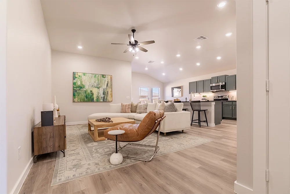 Living room with white sectional sofa, leather lounge chair, and wooden coffee table, open to kitchen with gray cabinets and barstool, ceiling fan, and multiple recessed lights.