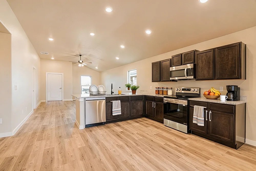 Open kitchen with dark wood cabinets, stainless steel appliances, light granite countertops, and light wood flooring, with a dining area and living space in the background.