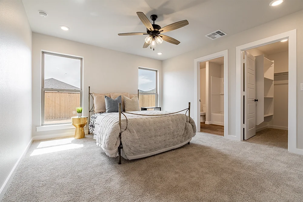 A bright bedroom with a bed, decorative pillows, a small gold side table with a plant, two windows, a ceiling fan, and an open closet with white shelves.