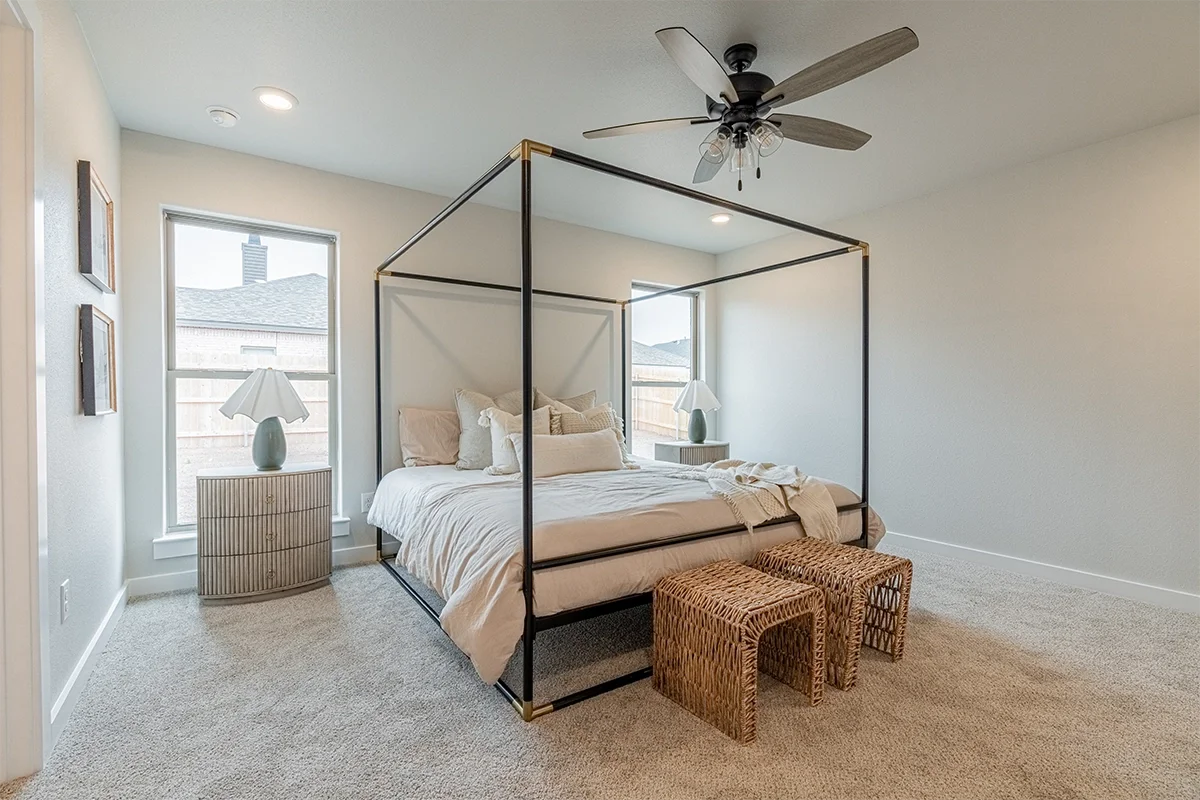 A bedroom with a four-poster bed, beige bedding, two nightstands with lamps, two small woven stools, and a ceiling fan.