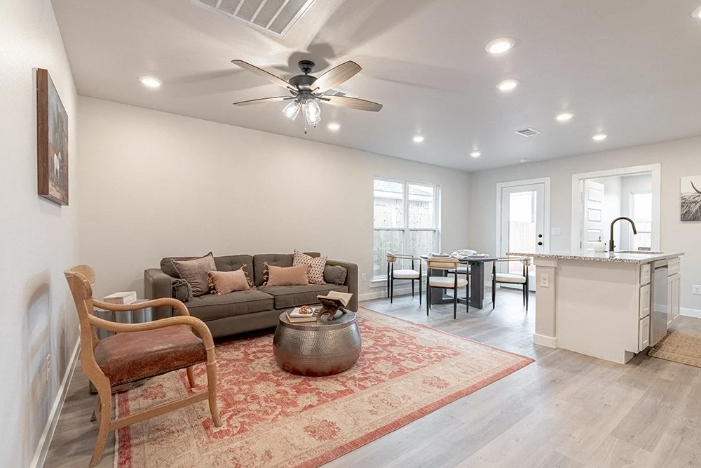 Living room with gray sofa, pink and beige pillows, wooden armchair, round metallic coffee table, area rug, dining table with chairs, kitchen island with sink, and windows with natural light.
