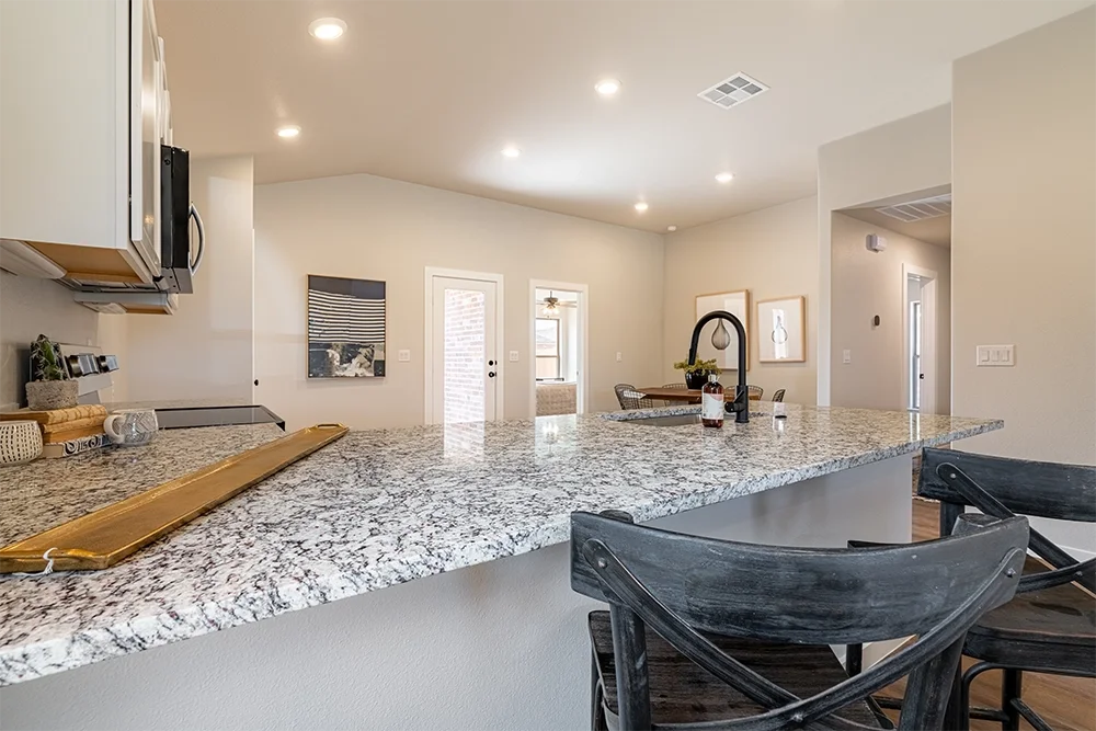 Modern kitchen with granite countertop, black chairs, and light-colored walls, with a view into the living or dining area.