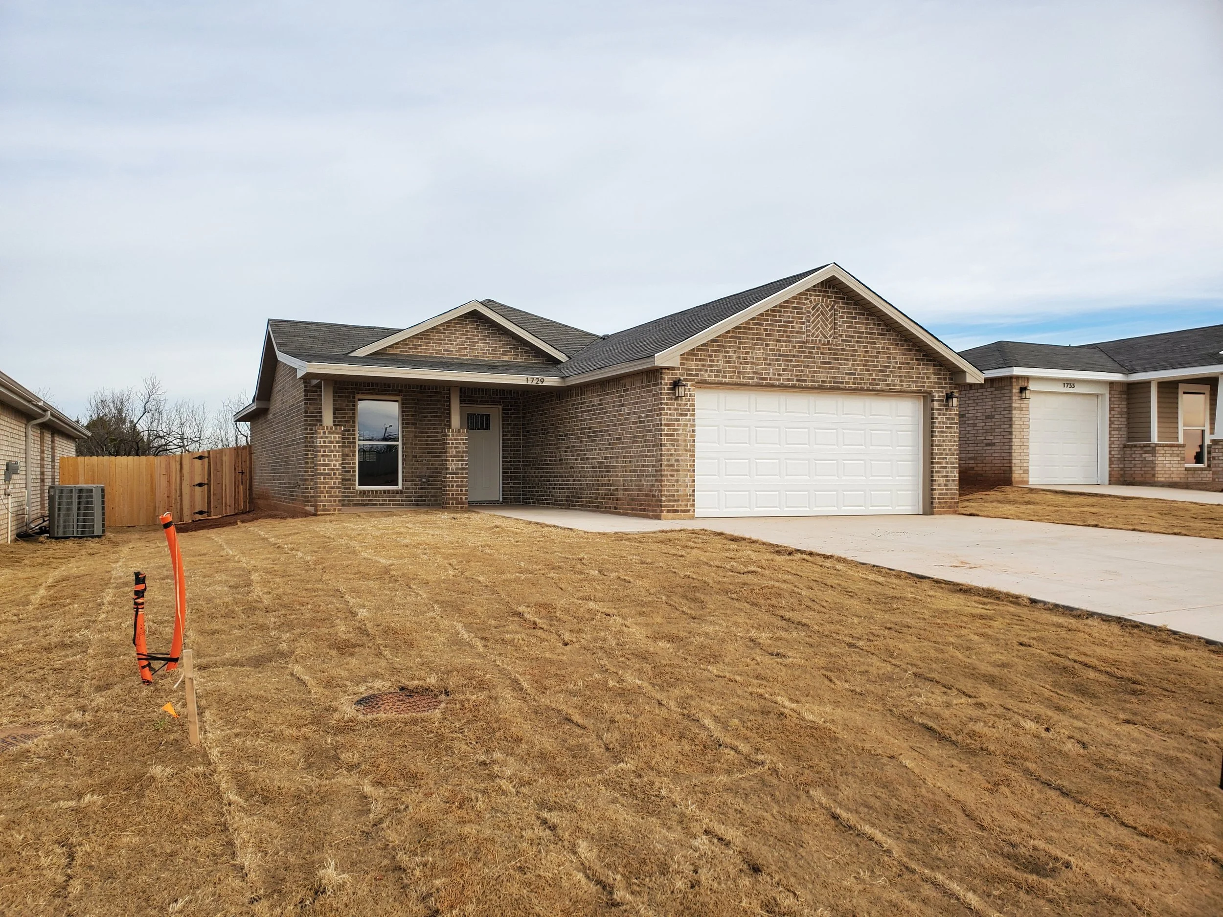 Front view of a newly built brick house with a white garage door, small front porch, and a front yard with patchy grass and some construction markers. The sky is clear with some clouds.