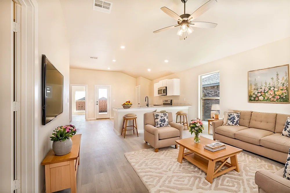 Bright open living room with beige sofas, a wooden coffee table, floral cushions, a TV mounted on the wall, and a kitchen in the background. Decor includes potted flowers and a wall art with flowers.