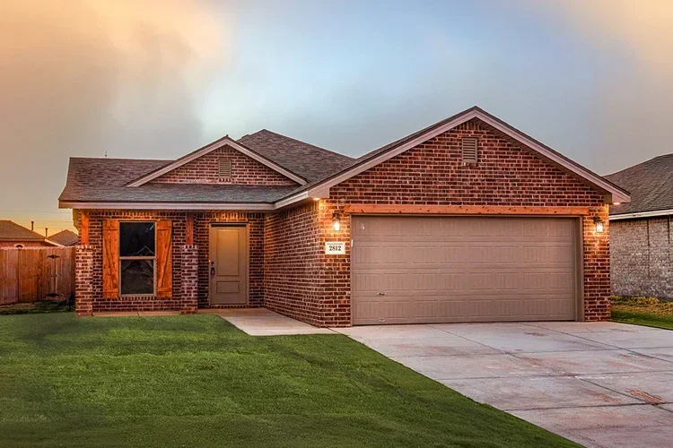 A single-story brick house with a two-car garage, front door, small front lawn, and a driveway at sunset.