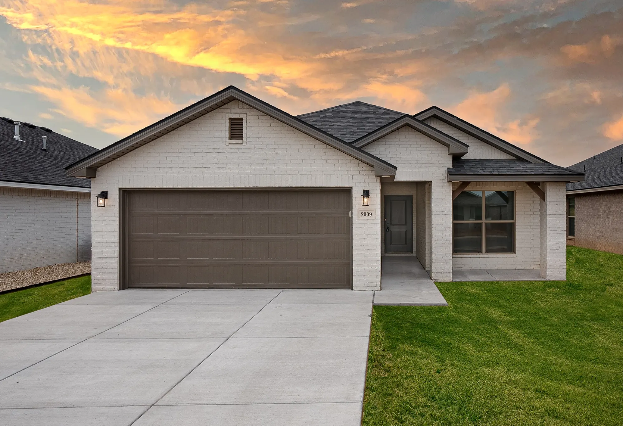 Front view of a modern white brick house with a brown garage door and front door, green lawn, and a sidewalk, during sunset with a colorful sky.