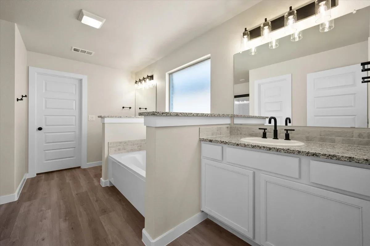 Modern bathroom with white cabinets, granite countertops, a large mirror, black fixtures, and a bathtub, featuring a wood floor and a frosted window.
