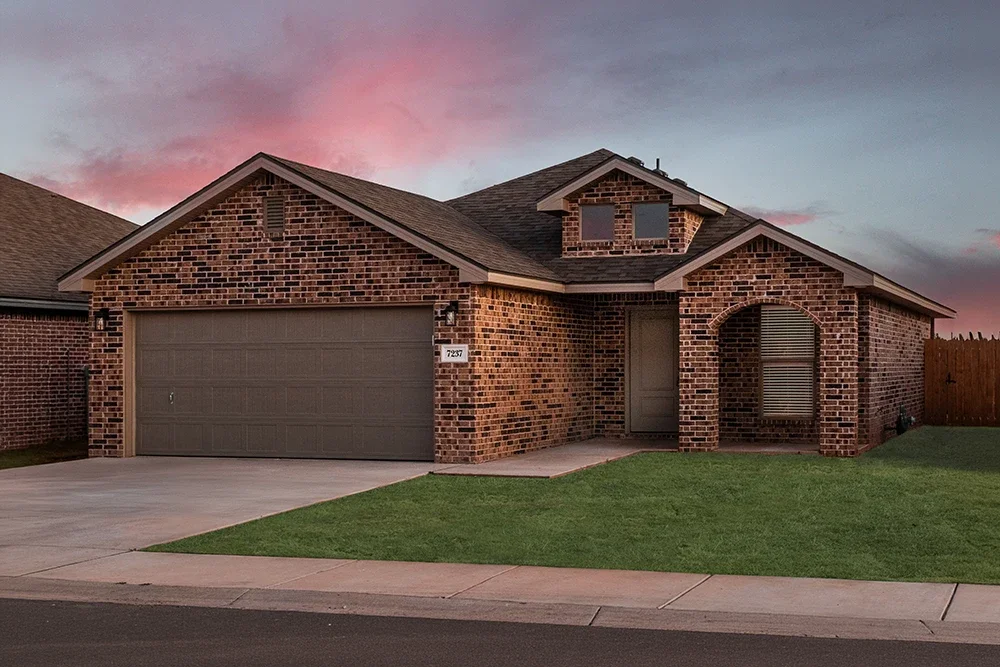 A brick suburban house with a two-car garage, a small front porch, and a well-maintained lawn during sunset.