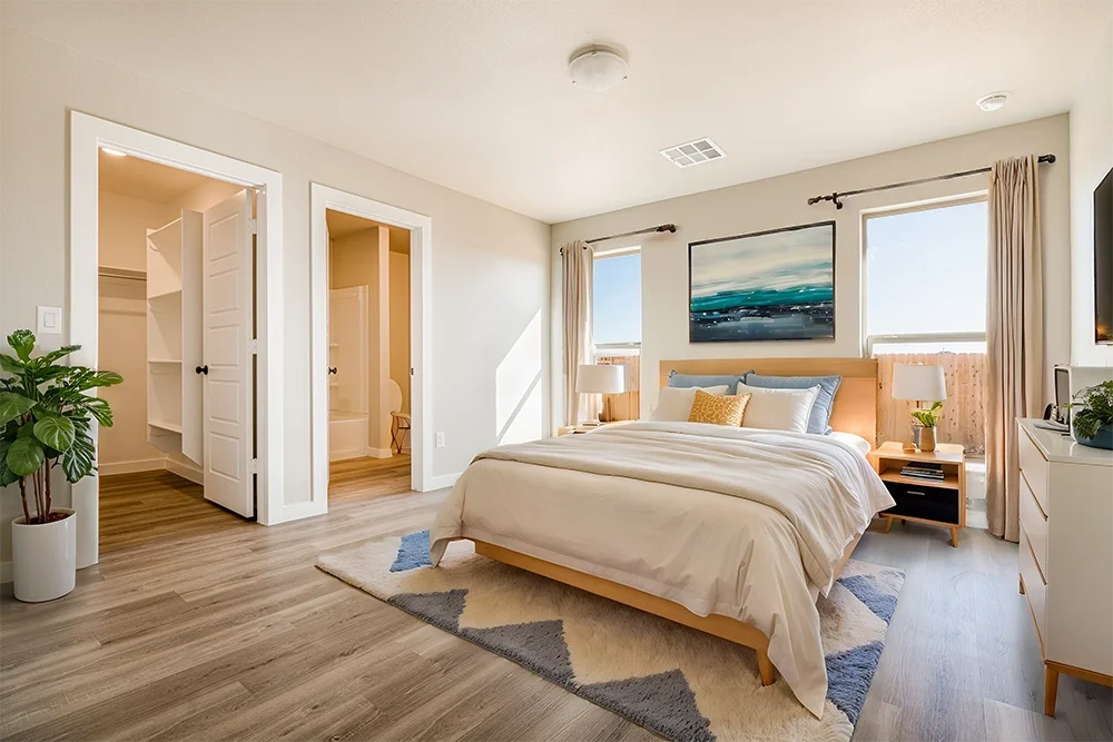 Bright bedroom with a neatly made bed, side tables with lamps, large windows with curtains, a painting above the bed, and a walk-in closet and bathroom visible through open doors.