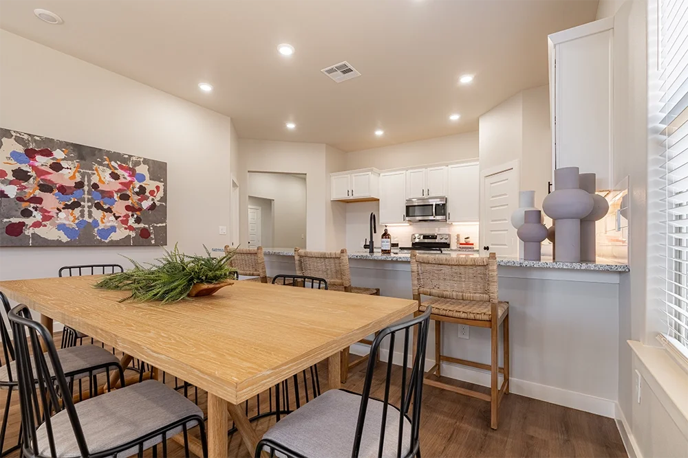 A dining area with a wooden table surrounded by black metal chairs and three woven-cane barstools at the kitchen counter. There is a large abstract painting on the wall and decorative vases on the counter. The kitchen has white cabinets, a microwave,