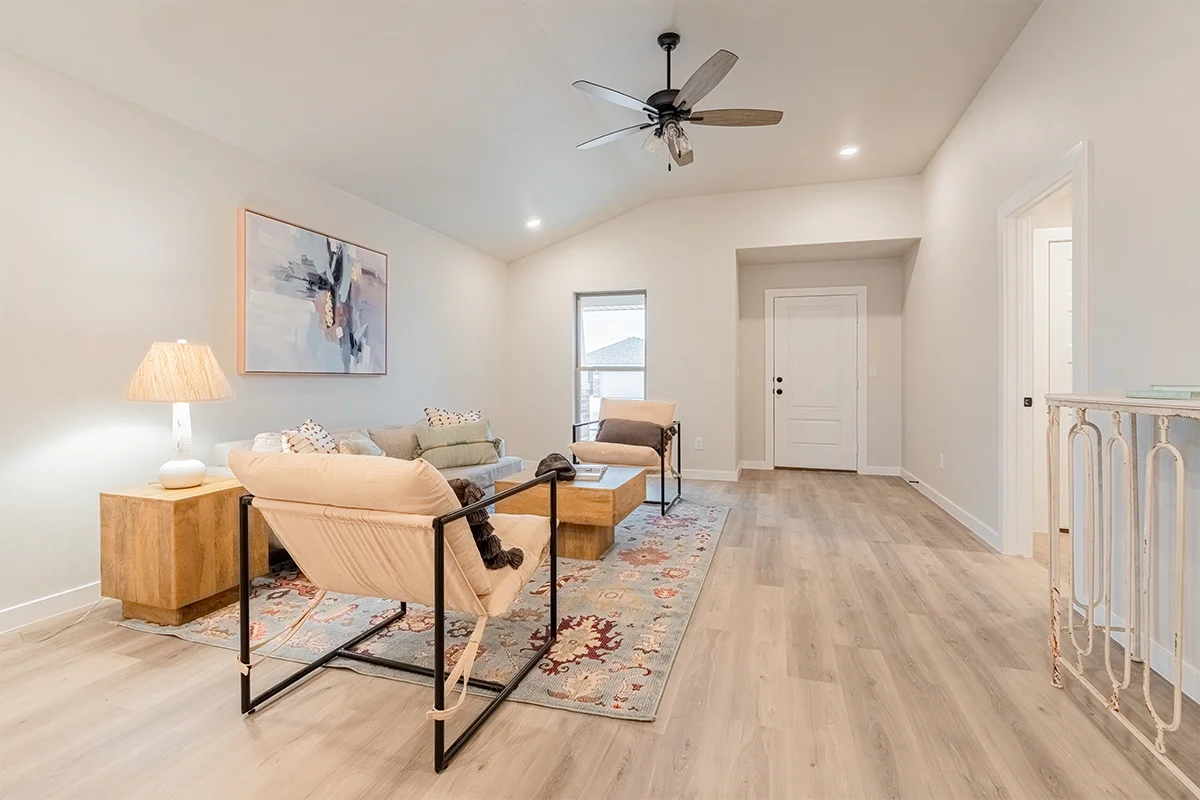 Living room with modern furniture, a wooden side table with a lamp, a white armchair, a beige sofa, a small wooden coffee table, an abstract painting, a ceiling fan, and hardwood floors.
