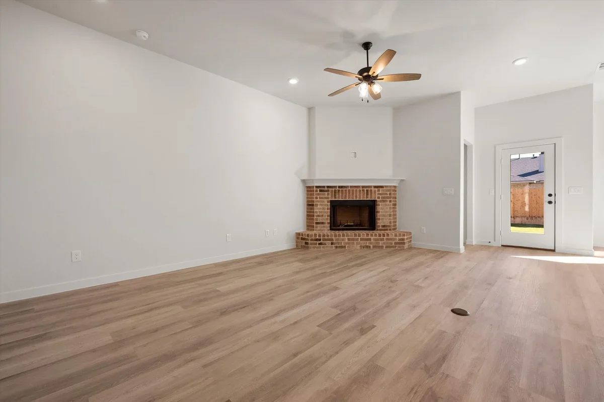 Empty living room with white walls, a brick fireplace, light wood flooring, a ceiling fan, and a glass door leading outside.
