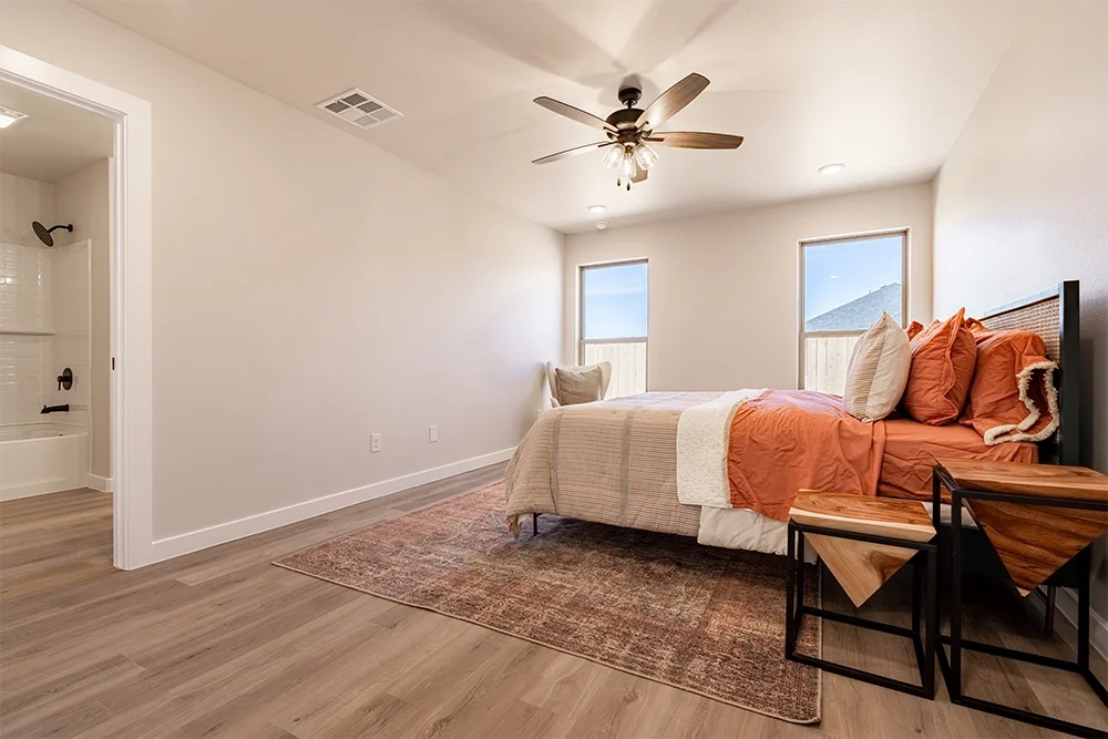 A bedroom with two windows, a bed with peach and white bedding, a wooden headboard, a ceiling fan, and a small area rug. There is a chair and side tables near the bed and an open bathroom door showing a shower.