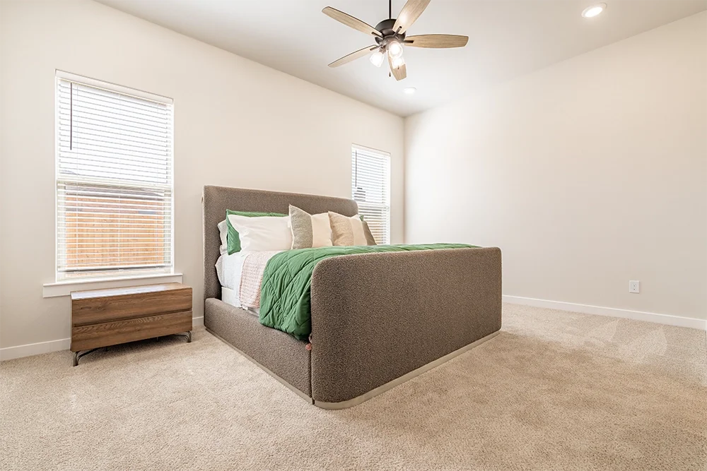 A bedroom with a gray upholstered bed, green bedding, and pillows, two windows with blinds, a wooden nightstand, a ceiling fan, and beige carpeting.