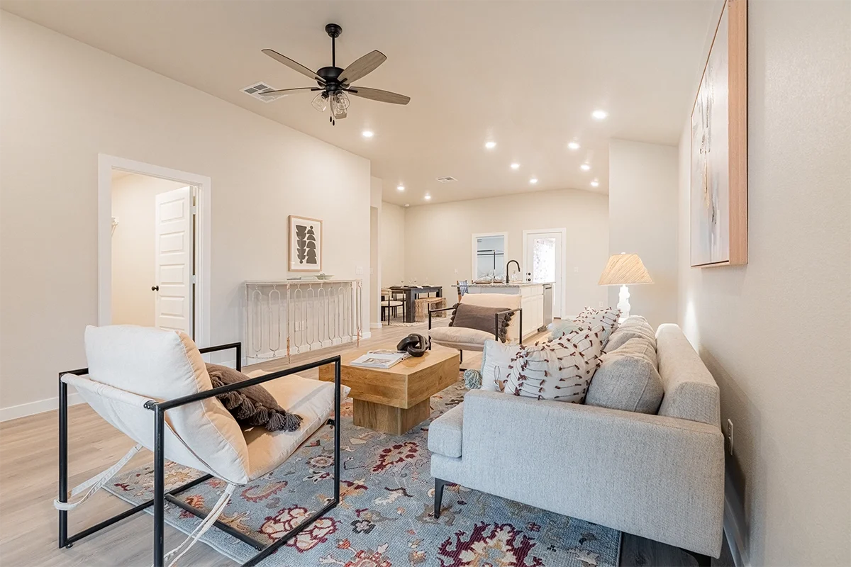 Living room with light-colored sofa, armchair with black metal frame, wooden coffee table, patterned area rug, and a wall with artwork. In the background, an open kitchen with a white island, and a dining area with chairs. Ceiling fan and recessed li