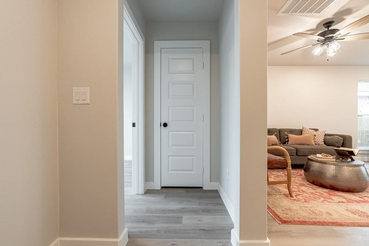 Interior view of a residential home showing a hallway with a white door and a living room with a gray sofa, patterned pillows, a round metallic coffee table, an area rug, and a ceiling fan with lights.