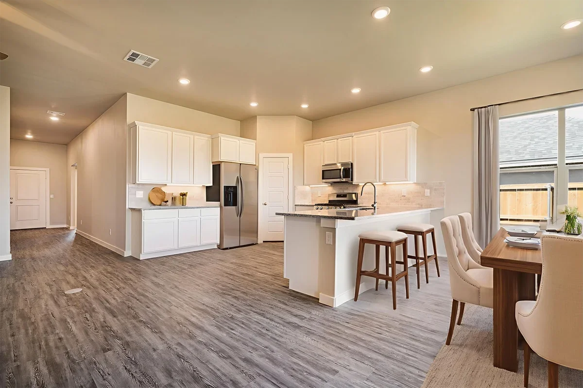 Open-concept kitchen with white cabinets, stainless steel appliances, a breakfast bar with two wooden stools, and a dining area with a wooden table and beige upholstered chairs. Large window with curtains and wood-look flooring.