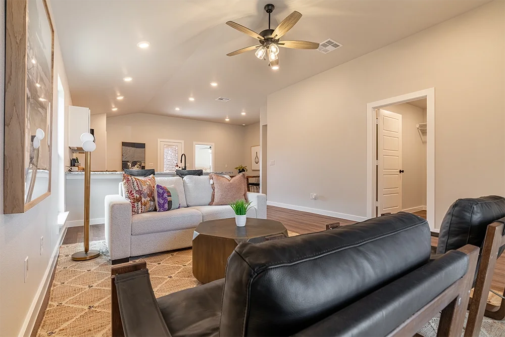 Living room with a beige sofa, black leather chairs, a wooden coffee table with a small plant, a white wall, a ceiling fan, and a doorway to a closet.