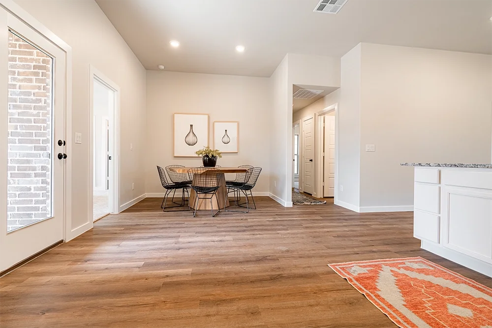 Empty dining area with a round wooden table, six wire chairs, two framed art pieces of vases on the wall, a door with brick design outside, and an entryway with white panel doors in the background. Hardwood floor with a patterned orange and white rug