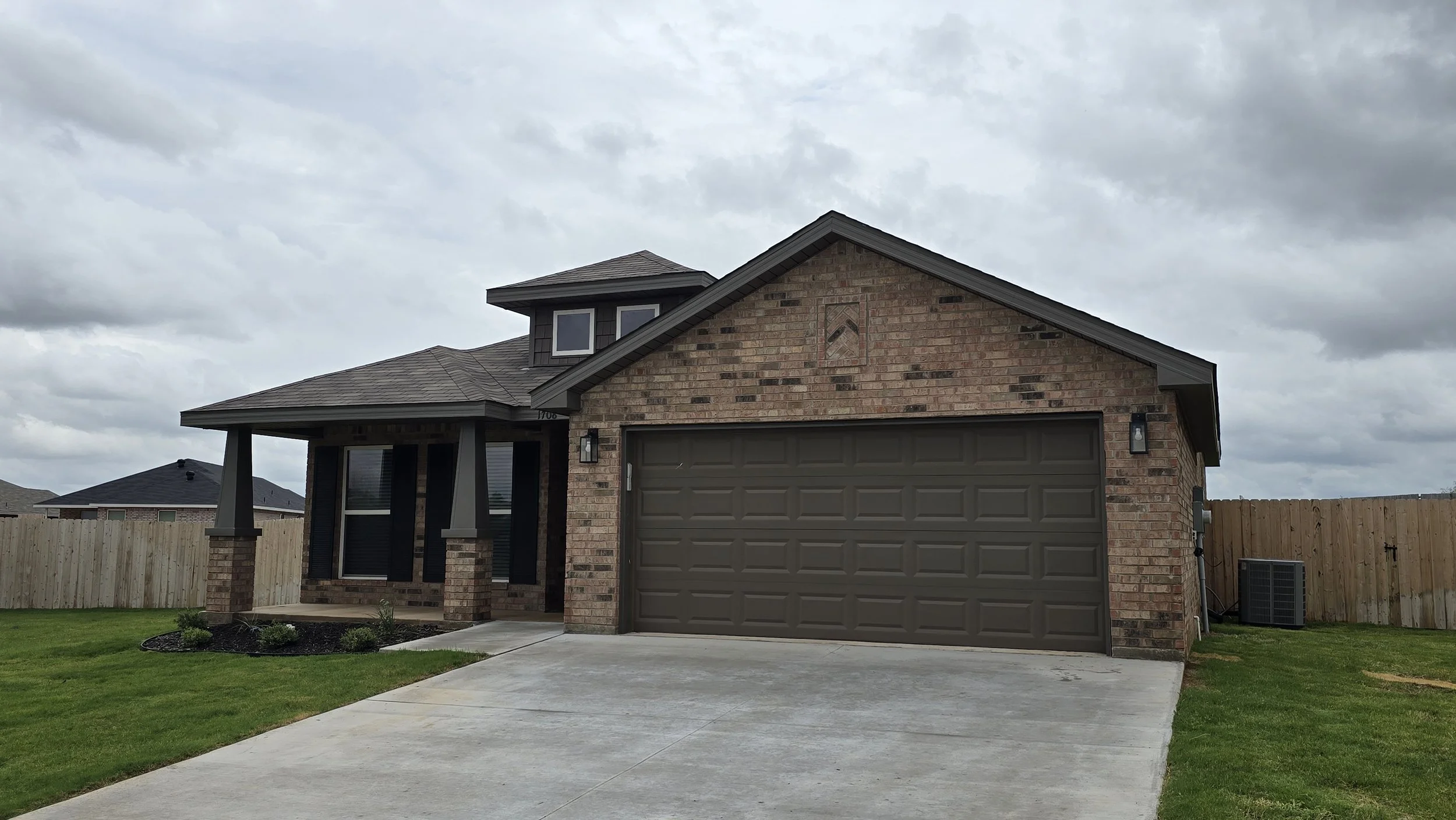 A suburban house with a brick exterior, attached garage, small front porch, a neatly manicured lawn, and a wooden fence in the backyard, under a cloudy sky.