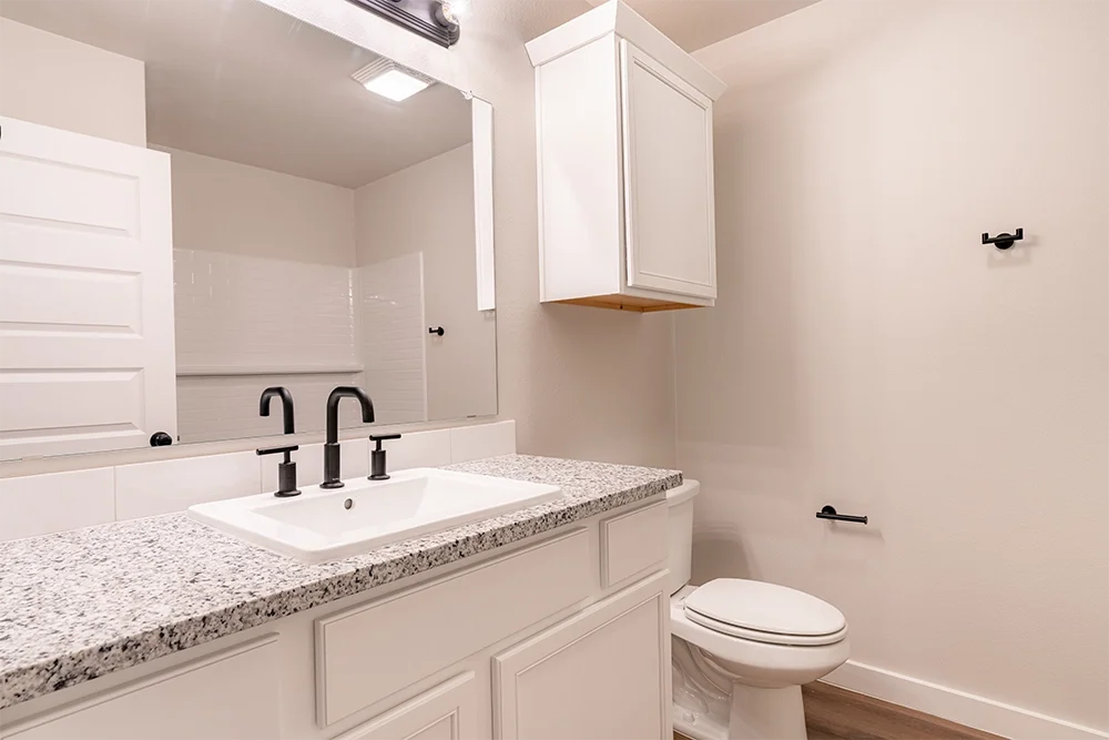 A small, modern bathroom with a granite countertop, white sink, black fixtures, and a white cabinet above the toilet.