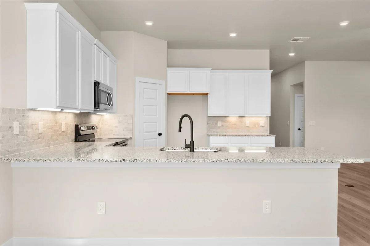 Modern kitchen with white cabinets, granite countertop island, stainless steel microwave, and stove, beige backsplash, and black faucet.