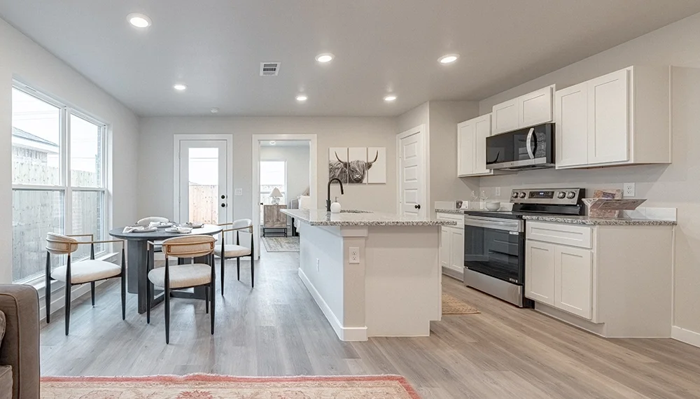 Open-concept kitchen and dining area with white cabinets, stainless steel appliances, granite countertops, a small island, a round dining table with four chairs, large windows, and a sliding glass door leading outside.