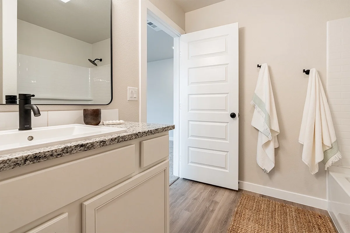 Bathroom with a granite countertop, white cabinet, black faucet, large mirror, towels on black hooks, and a white door slightly open to another room.