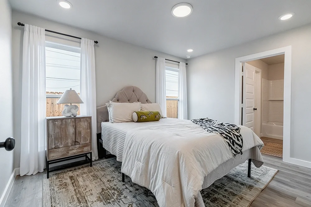 Bedroom with a bed, nightstand, lamp, windows with white curtains, and an adjoining bathroom.