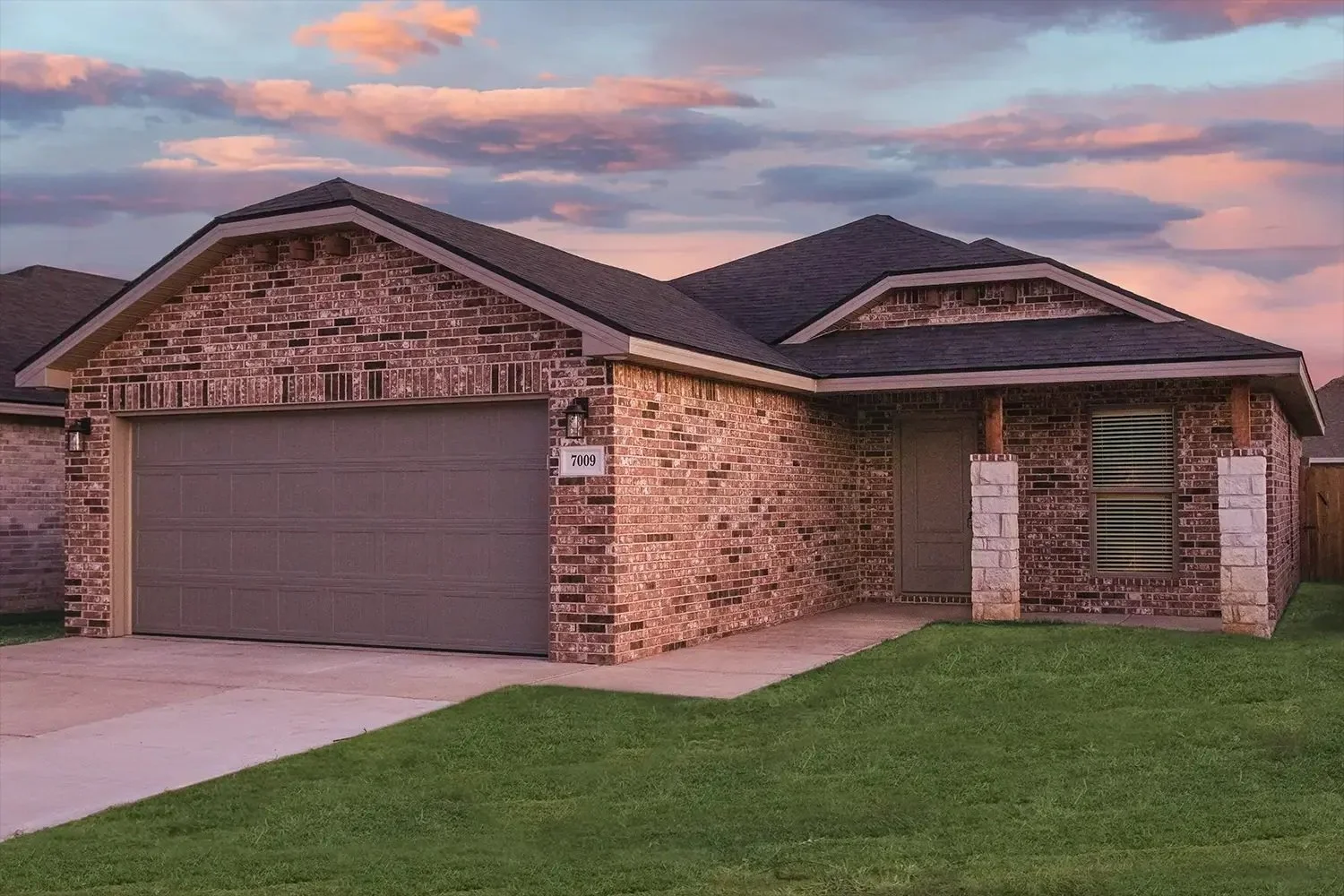 A brick house with a two-car garage, green lawn, and a sunset sky with pink and blue clouds.
