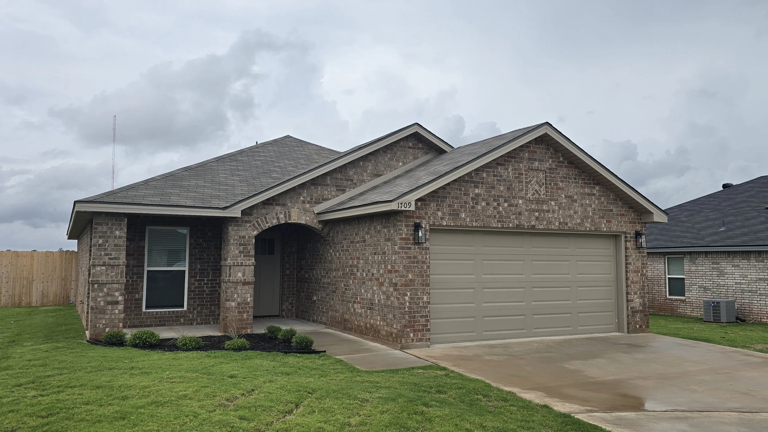 A brick house with a gray garage door and a front porch with a small garden, under an overcast sky.
