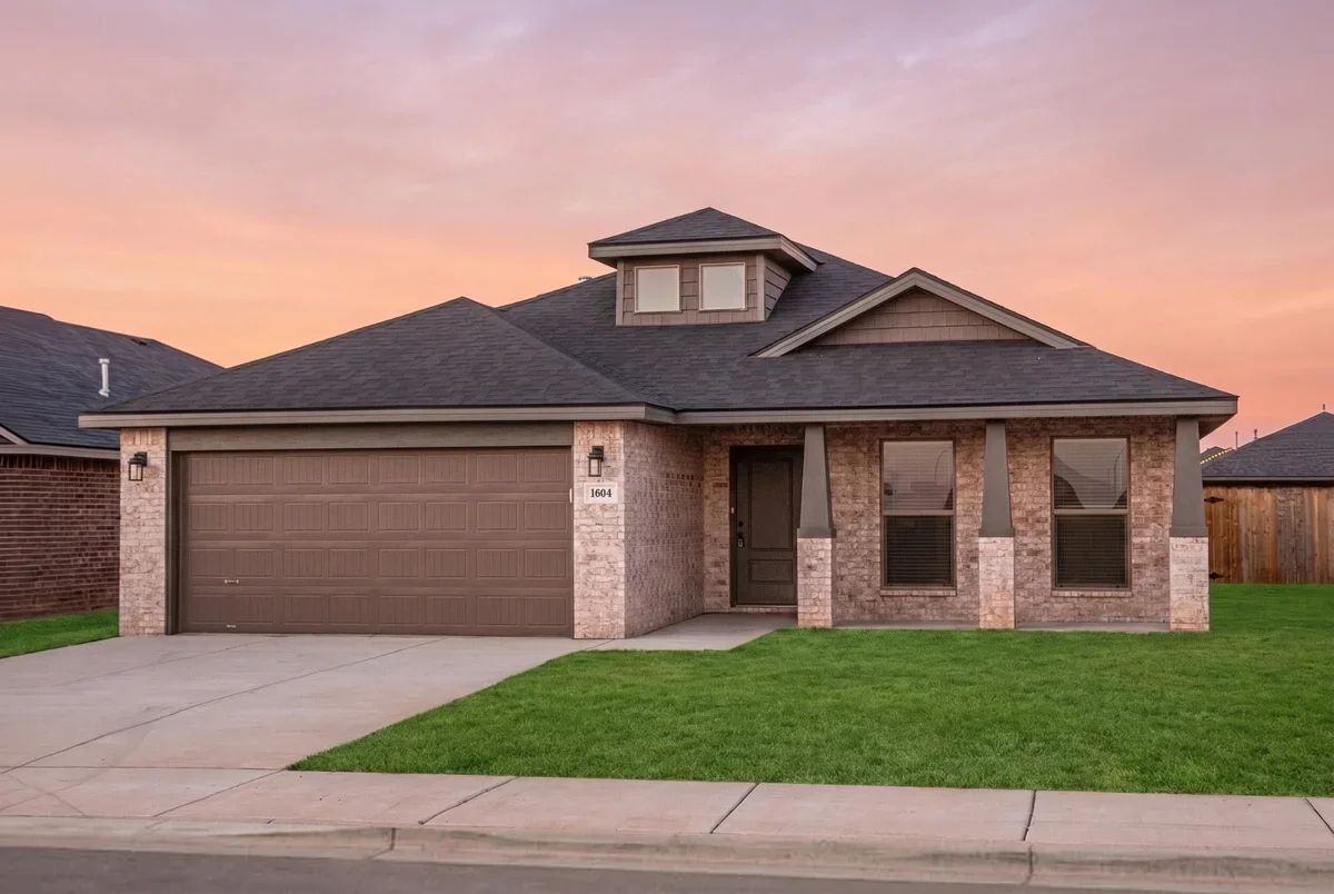 A new brick house with a two-car garage, dark roof, small front yard, and pinkish sunset sky in the background.