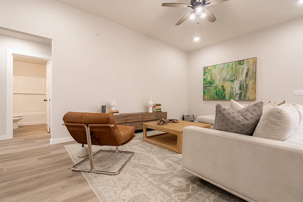 Living room with white walls, a white sofa with gray pillows, a wooden coffee table, a brown leather chair, a wooden TV stand with decorative items, a colorful abstract painting on the wall, a ceiling fan, and an adjacent bathroom with visible toilet