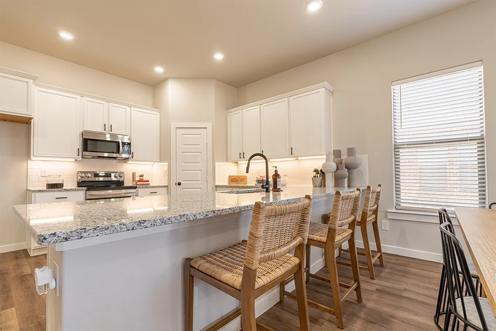 Modern kitchen with white cabinets, granite countertops, stainless steel appliances, a breakfast bar with wicker chairs, and a large window with blinds.