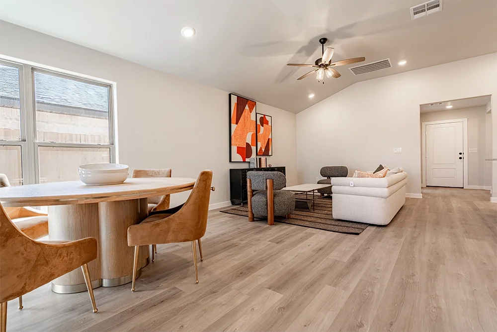 Open living and dining area with beige walls, wood flooring, and modern furniture. The dining table has four tan chairs, and the living room has a white couch, gray accent chairs, a coffee table, and a black sideboard with a geometric artwork above i