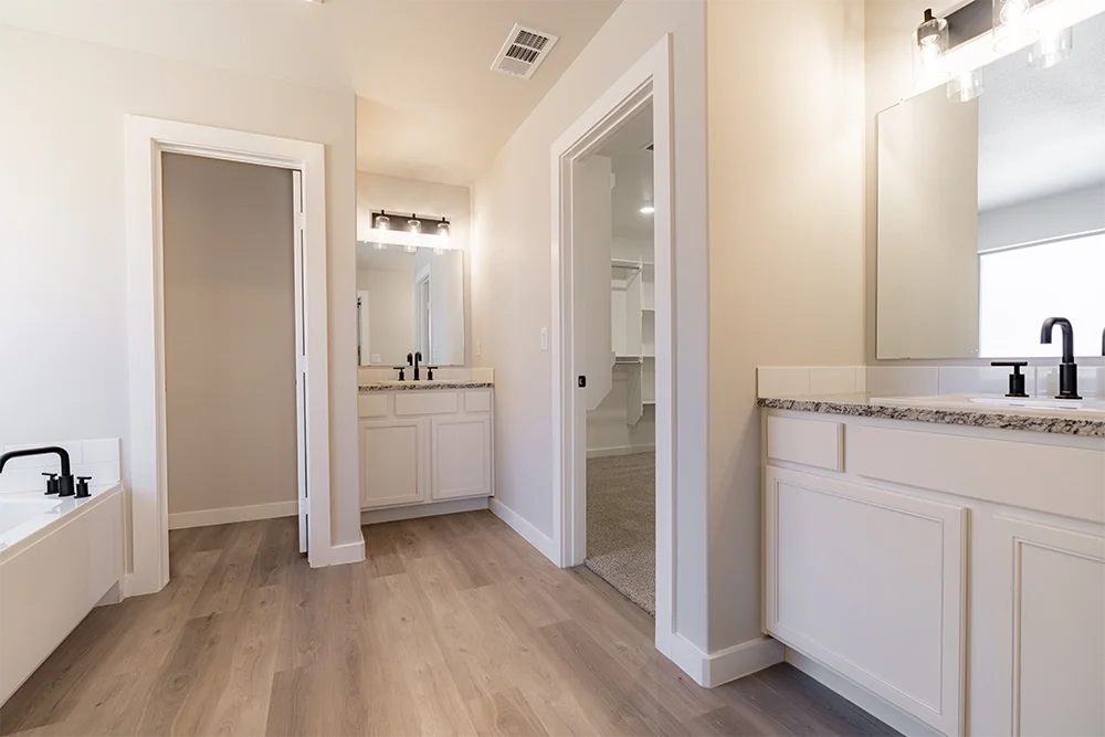 A spacious bathroom with a bathtub on the left, a vanity with mirror and lighting in the middle, and a mirror and sink on the right, featuring white cabinetry and granite countertops.