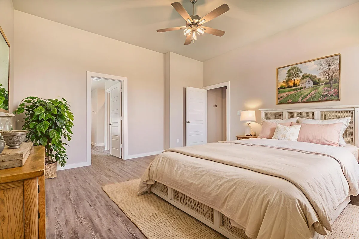 A bedroom with a large bed, light-colored bedding, a nightstand with a lamp, a framed landscape painting above the headboard, a ceiling fan, and a potted plant near the door.