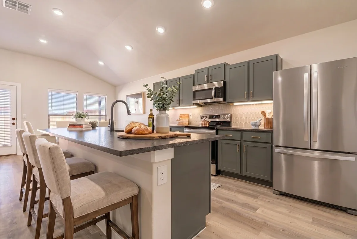 Modern kitchen with grey cabinets, stainless steel refrigerator, oven, and microwave, island with beige chairs, natural light, and a wooden floor.