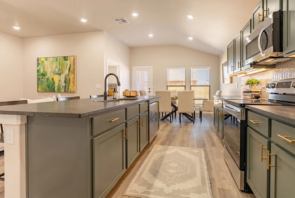 Modern kitchen with green cabinets, gray countertops, and gold hardware, open to a dining area with chairs and windows.