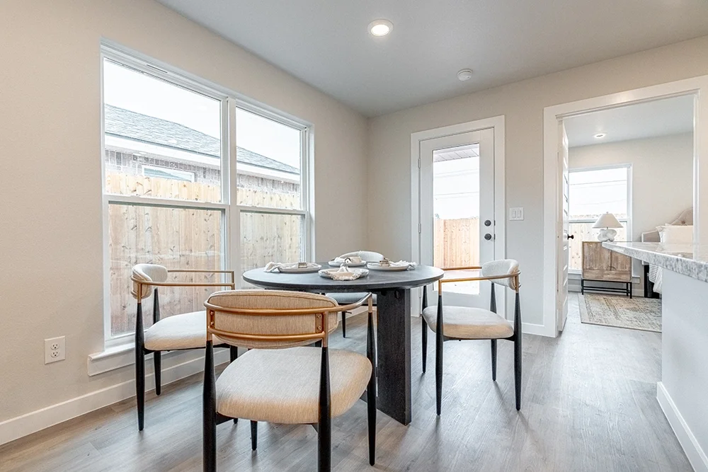 A dining area with a round black table set with plates and napkins, surrounded by five beige chairs with black legs, near large windows and a door leading outside, with a living room visible in the background.