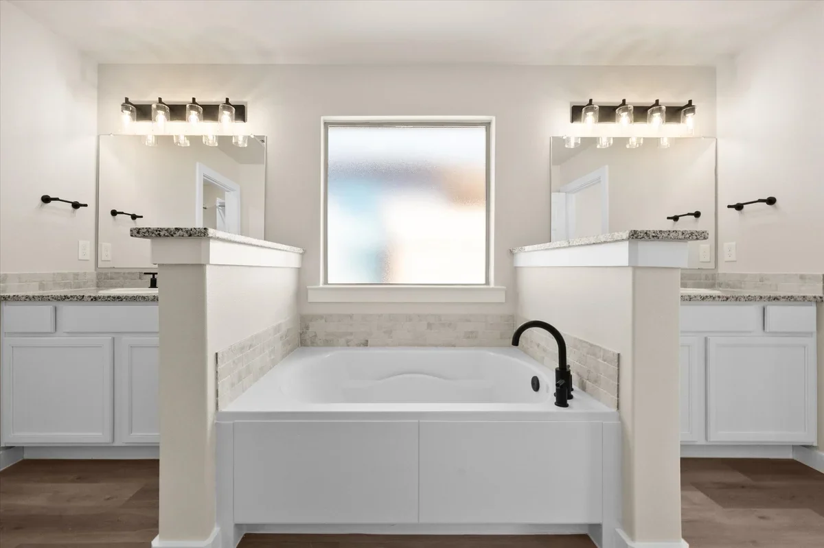 Bathroom with a central bathtub, two vanities with granite countertops and large mirrors, black fixtures, and a frosted window above the tub.
