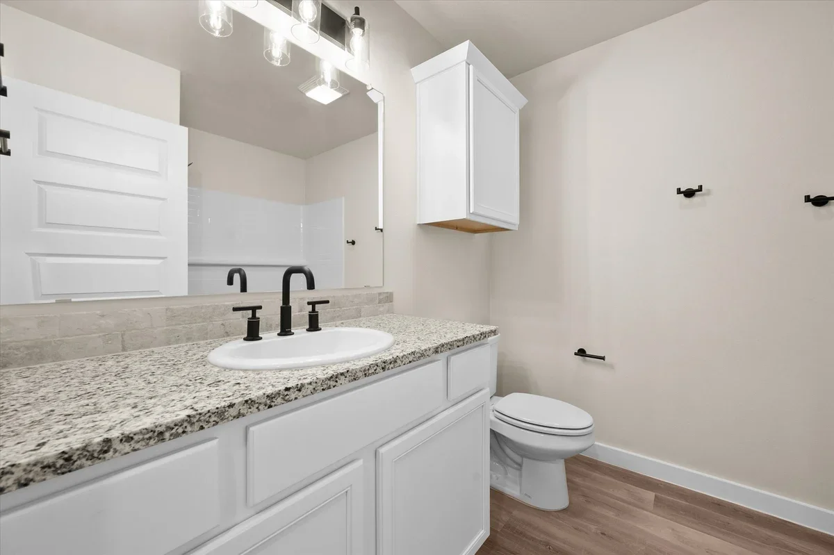 A modern bathroom with a granite countertop sink, black fixtures, white cabinets, a white toilet, a wall-mounted cabinet, and a large mirror with ceiling light fixtures.