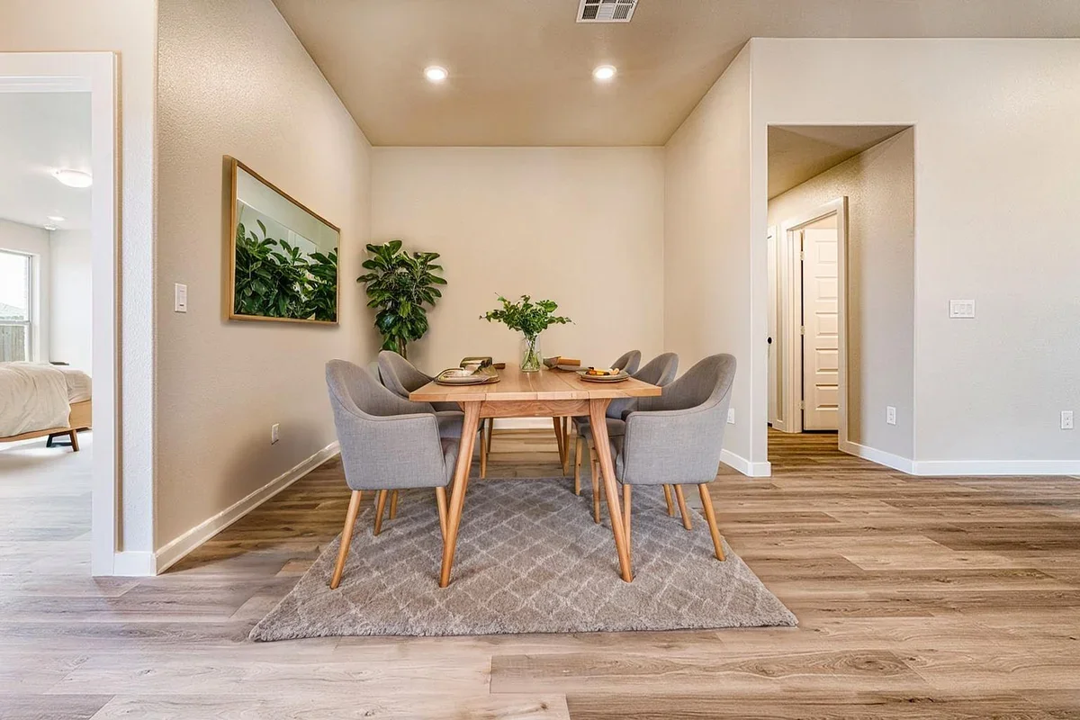 Dining room with a wooden table surrounded by six gray upholstered chairs, a vase with green leaves on the table, a large mirror on the wall, and two tall plants in the corner.