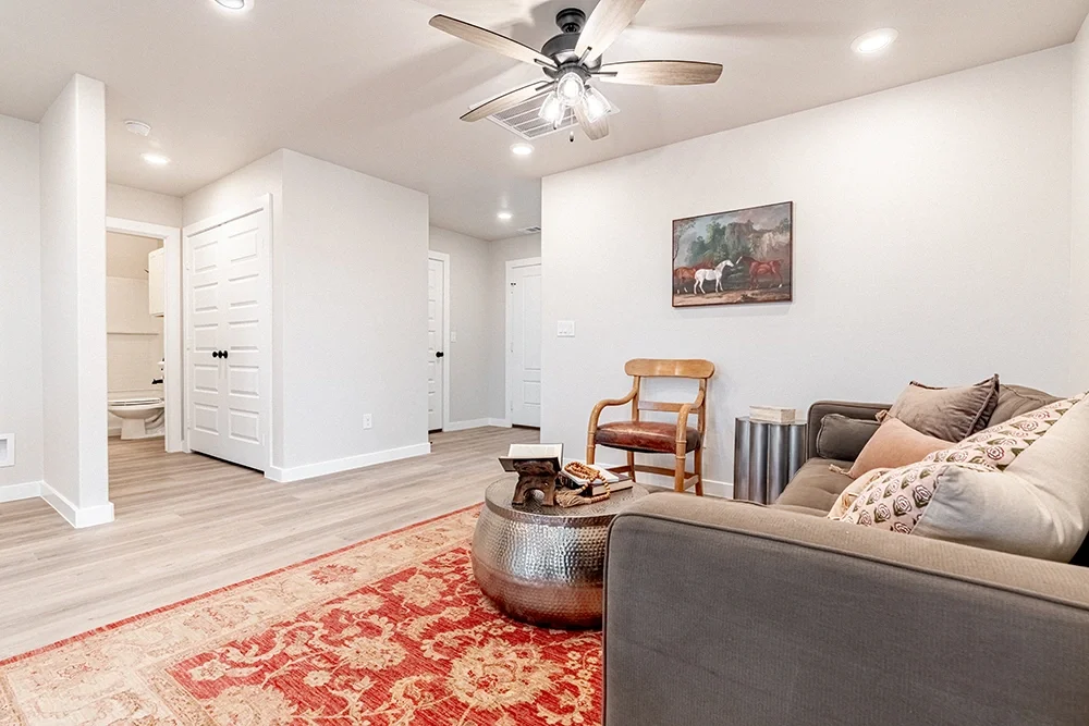 Living room with a beige sofa, pillows, a round metallic coffee table, a wooden chair, a framed painting of horses on the wall, and a ceiling fan.