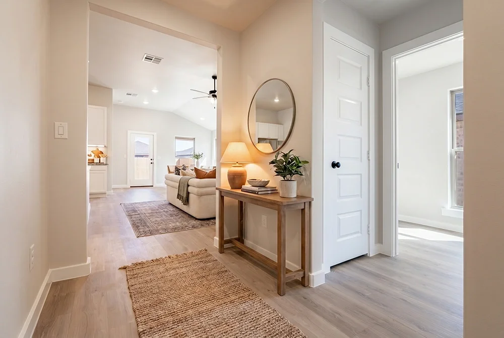 Bright living room and entryway in a modern home with light-colored walls, hardwood floors, a round mirror, a wooden console table with a lamp and plant, and a white couch in the background.